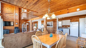 A cozy cabin interior with a wooden dining table, chairs, a sofa, a kitchen area, and a TV. - Cavallo Dunes - 4x4 Carova Beach, Oceanfront, Ocean View, Hot Tub, Pets OK, Year Round Flex Stays (4x4 - Carova Beach)