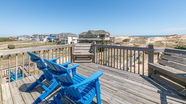 A wooden deck with blue Adirondack chairs, a wooden walkway, and a view of beach houses and the ocean. - Cavallo Dunes - 4x4 Carova Beach, Oceanfront, Ocean View, Hot Tub, Pets OK, Year Round Flex Stays (4x4 - Carova Beach)