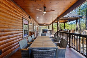Eine überdachte Terrasse im Freien mit einem langen Holztisch und Korbstühlen, umgeben von Holzgeländern und Blick auf Bäume.