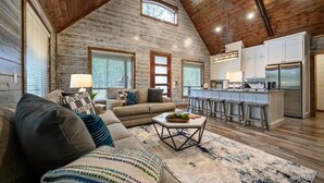 A spacious living room with a high ceiling and wooden beams, a cozy seating area with a sofa and cushions, a dining table with a fruit bowl, and a modern kitchen with white cabinets and stainless steel appliances. - 3 bedroom with game room! (Broken Bow)