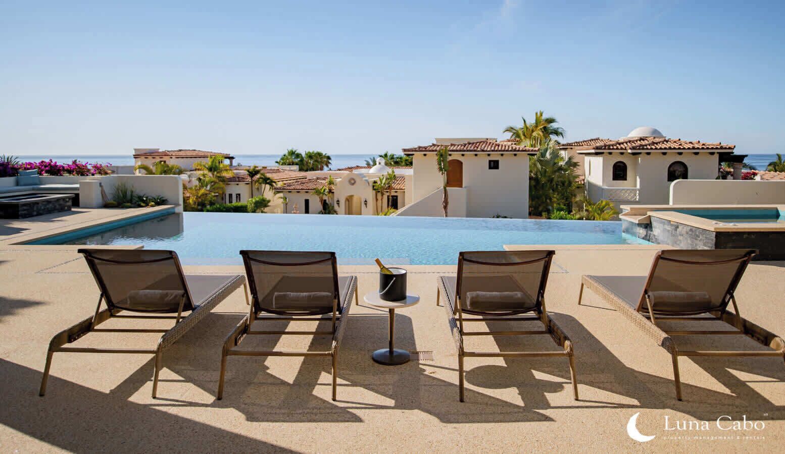 A poolside area with lounge chairs and a small table with a bottle, overlooking a pool and a coastal view.