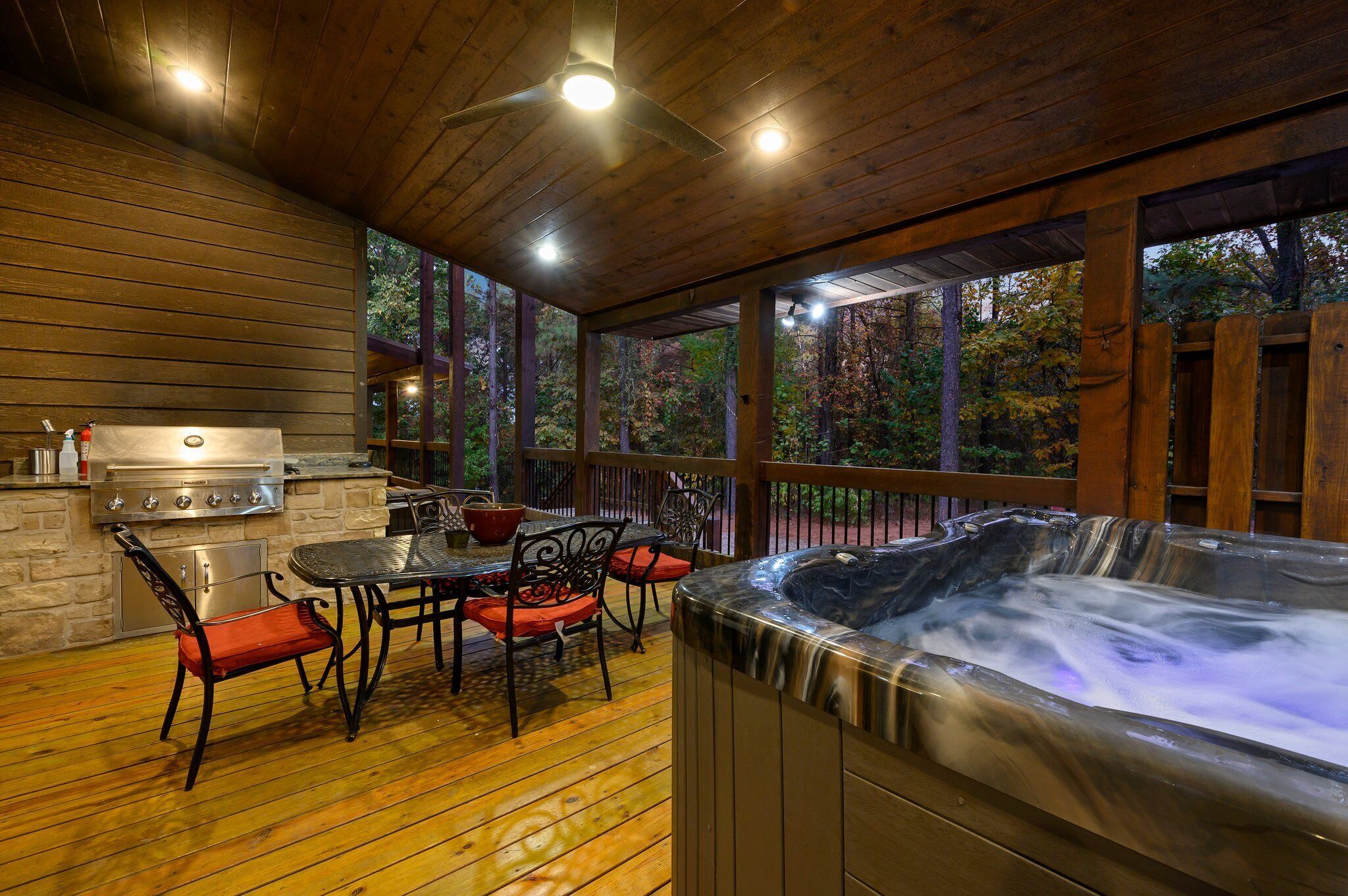A covered outdoor patio with a hot tub, a grill, and a dining area with chairs and a table.