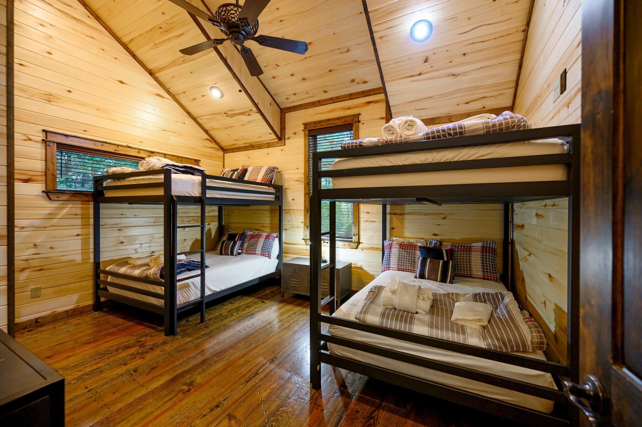 A cabin with two bunk beds, wooden walls, a ceiling fan, and a window with blinds.