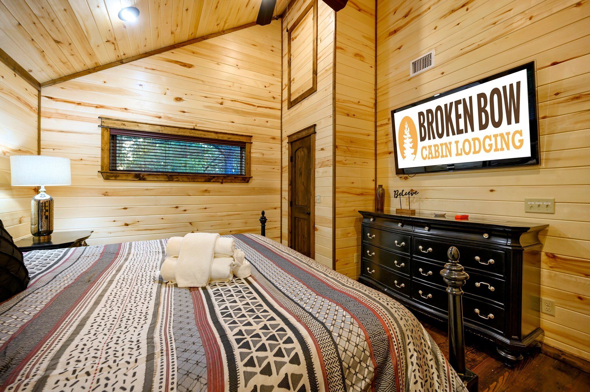 A cabin room with a bed, a dresser, a window, and a sign that reads "Broken Bow Cabin Lodging."