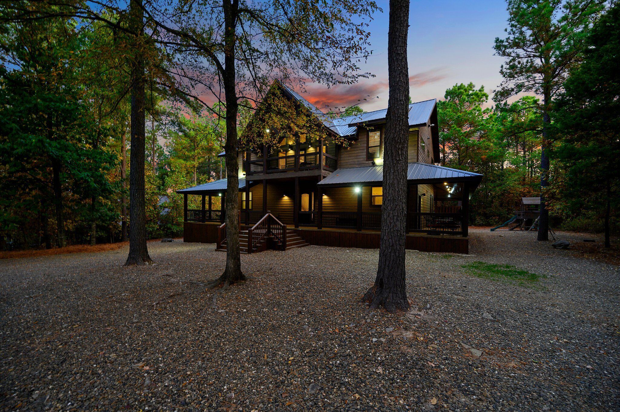 A large, multi-story wooden cabin surrounded by trees, with a porch and steps leading to the entrance.