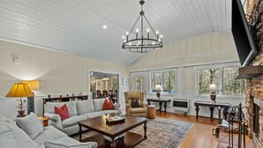 A spacious living room with a fireplace, a chandelier, and a large window providing a view of the outdoors. - Duck's Nest 3BR: Modern Creekside Retreat w/ Large Deck, Private Lower Suite (Highlands)