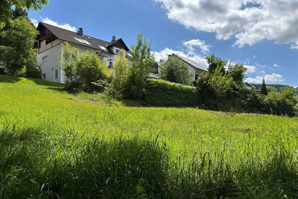 A two-story house with a sloped roof, surrounded by lush greenery and a grassy field. - Platz, Charme & ein Hauch Luxus: Happy Place mit Whirlpool, Sauna und Traumblick (Plettenberg)