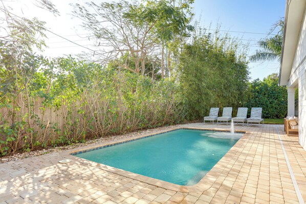 A rectangular swimming pool with a tiled edge, surrounded by a brick patio. There are lounge chairs and a small table with cushions.