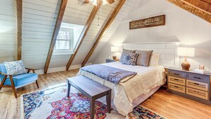 A bedroom with a sloped ceiling, wooden beams, a large bed, a nightstand, a chair, and a colorful rug. - Dog-friendly Hiawassee Cabin Near Lake & Hiking (Hiawassee)