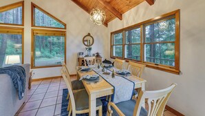Un coin repas avec une table en bois dressée pour quatre personnes, entourée de chaises, de grandes fenêtres donnant sur la forêt et un luminaire suspendu.