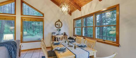 A dining area with a wooden table set for four, surrounded by chairs, large windows with forest views, and a hanging light fixture.