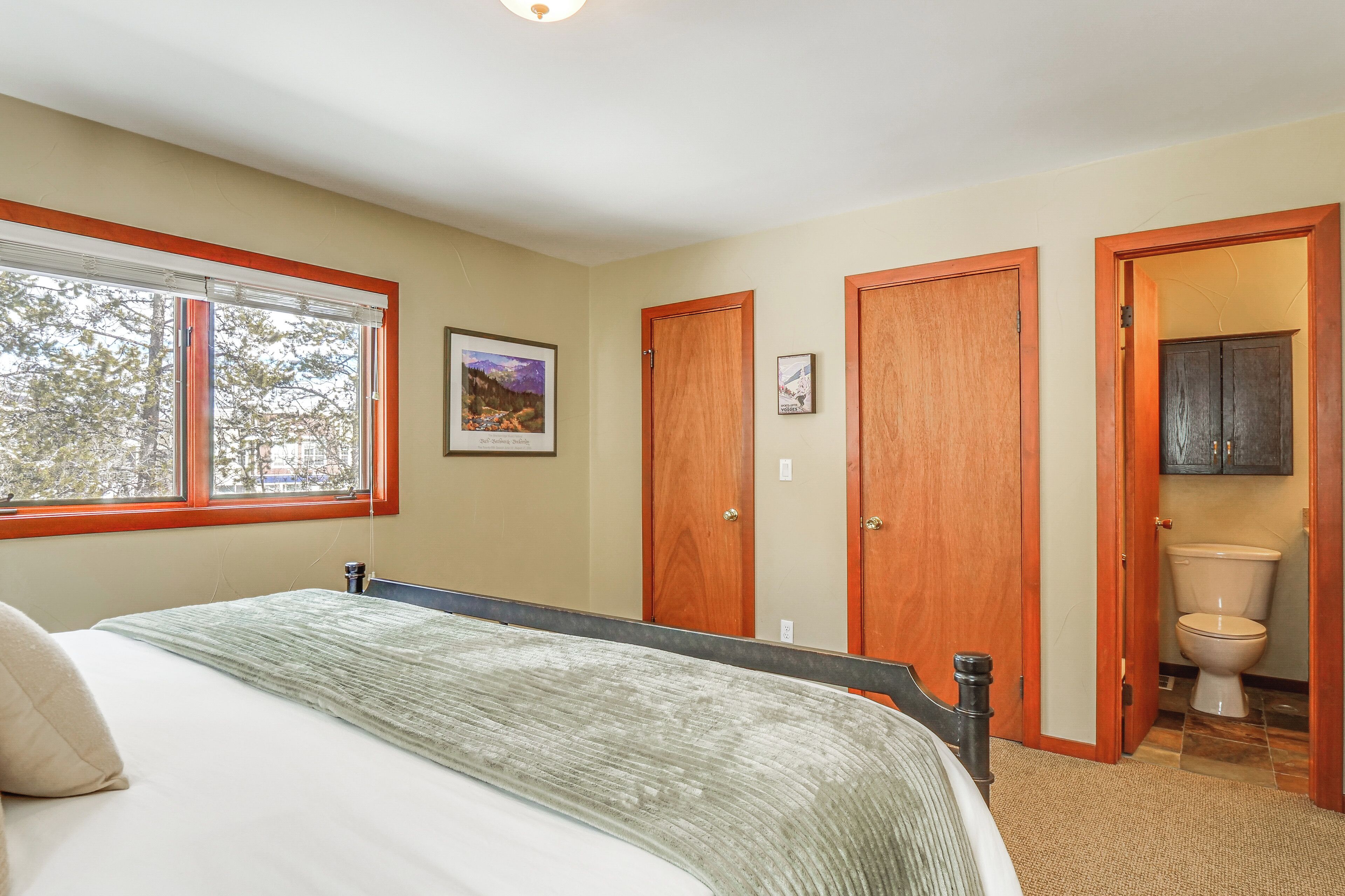 Une chambre à coucher avec un lit, une fenêtre, une porte et des toilettes visibles à travers une porte entrouverte.