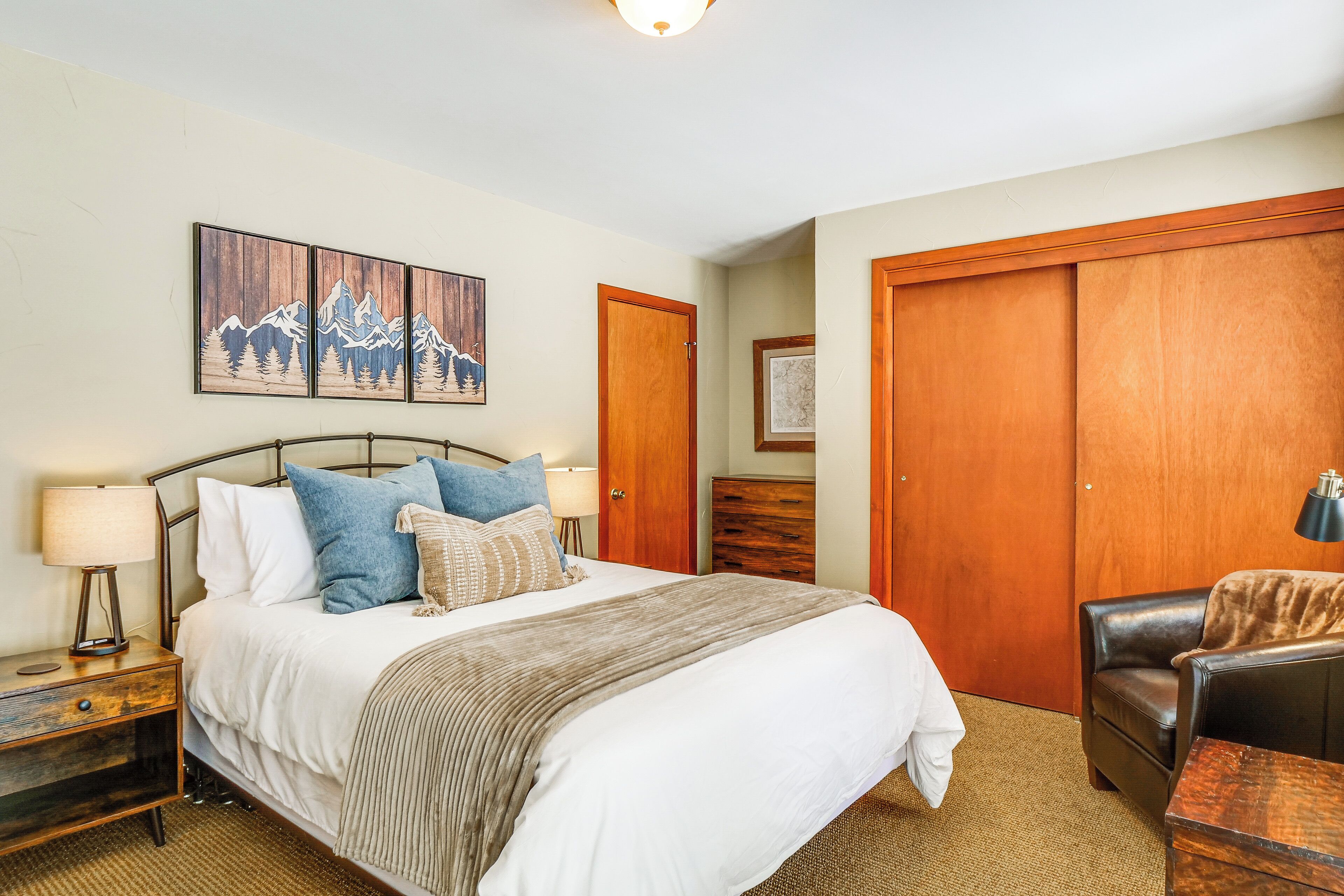 A neatly arranged bedroom with a large bed, a wooden wardrobe, a leather armchair, and a nightstand with a lamp.