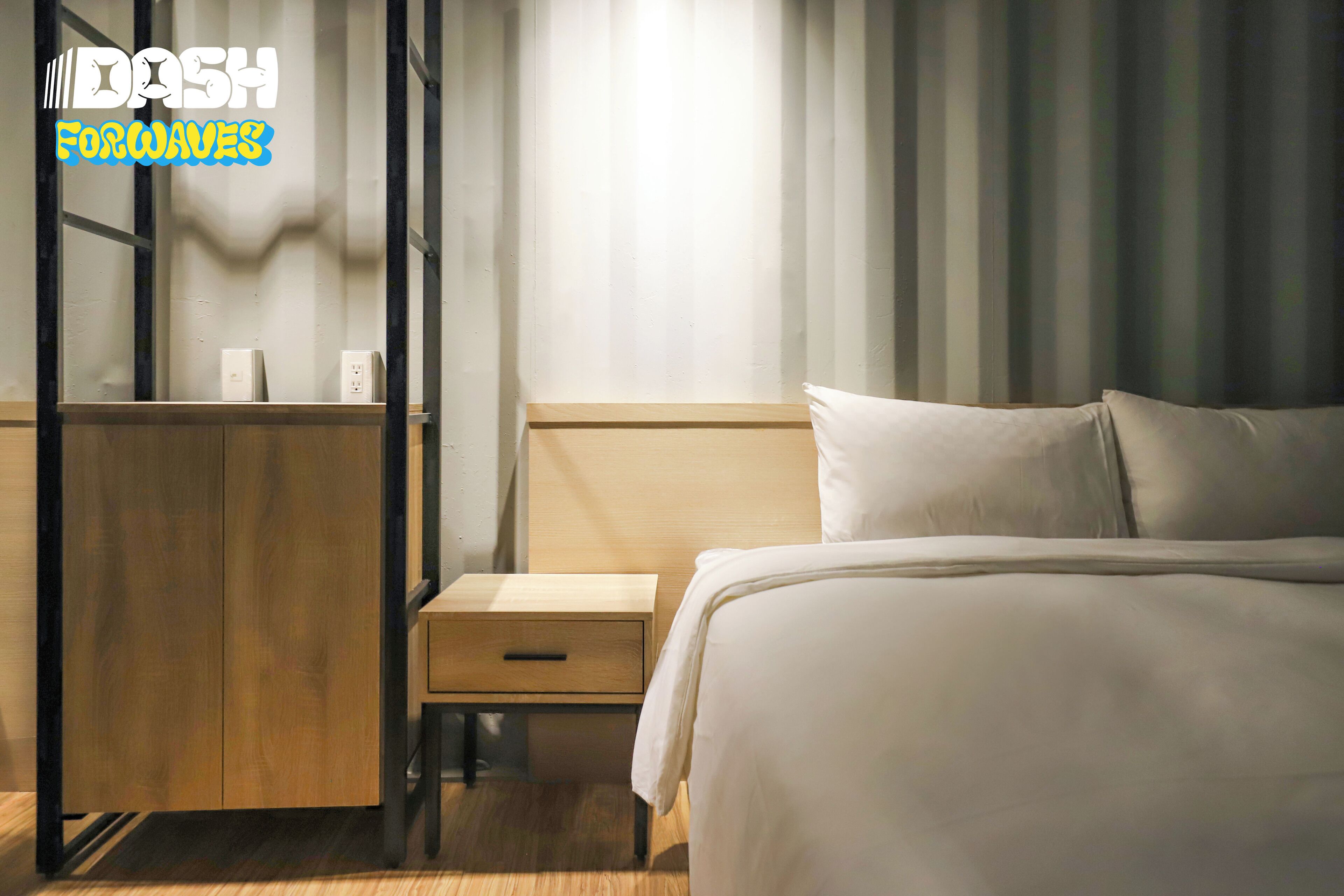 A hotel room with a wooden nightstand, a bed with white bedding, and a window with vertical blinds.