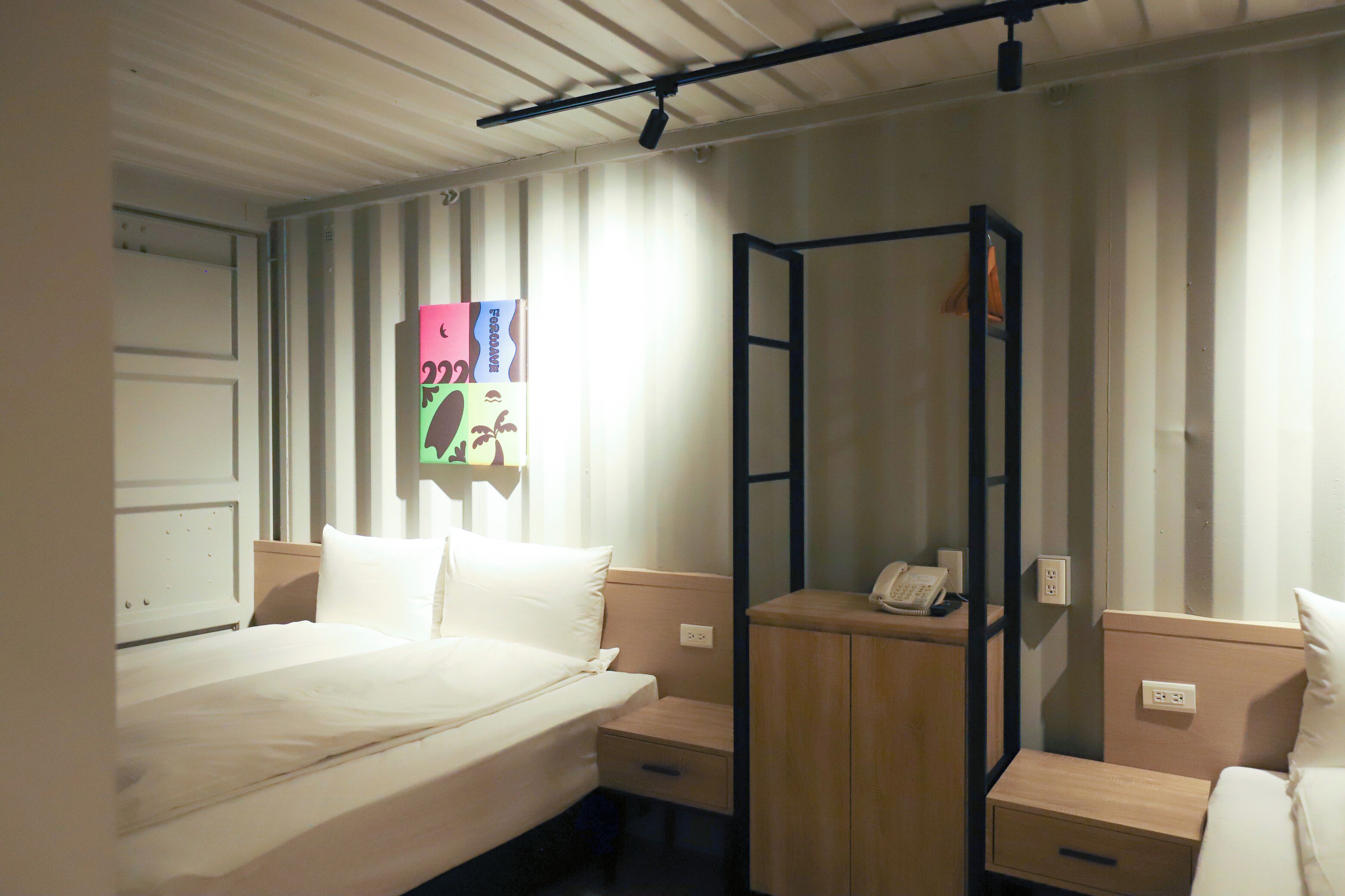 A hotel room with a bed, a bedside table with a phone, a small cabinet, and a wall-mounted shelf with a colorful item.