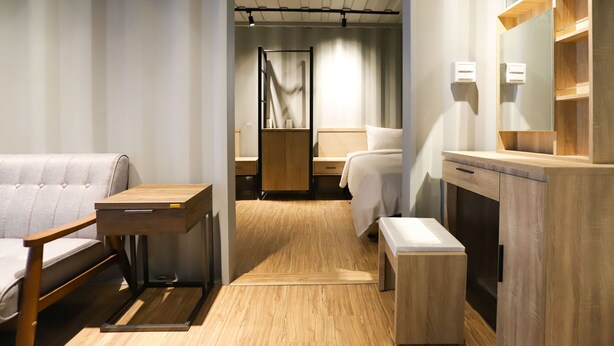 Una habitación de hotel moderna con piso de madera, una cama con colchón blanco, una mesita de noche de madera, un banco de madera, un escritorio de madera y un armario con espejo.