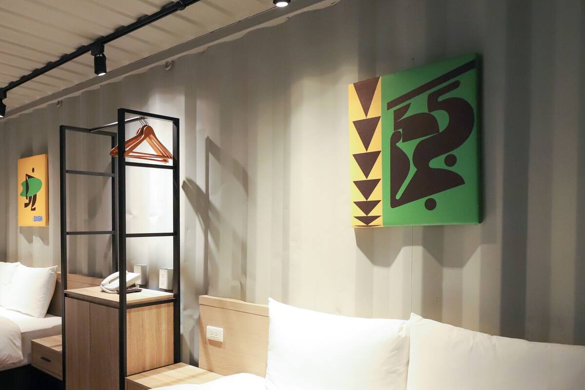 A hotel room with a bed, a wooden nightstand, a hanging coat rack, and a wall with geometric artwork.