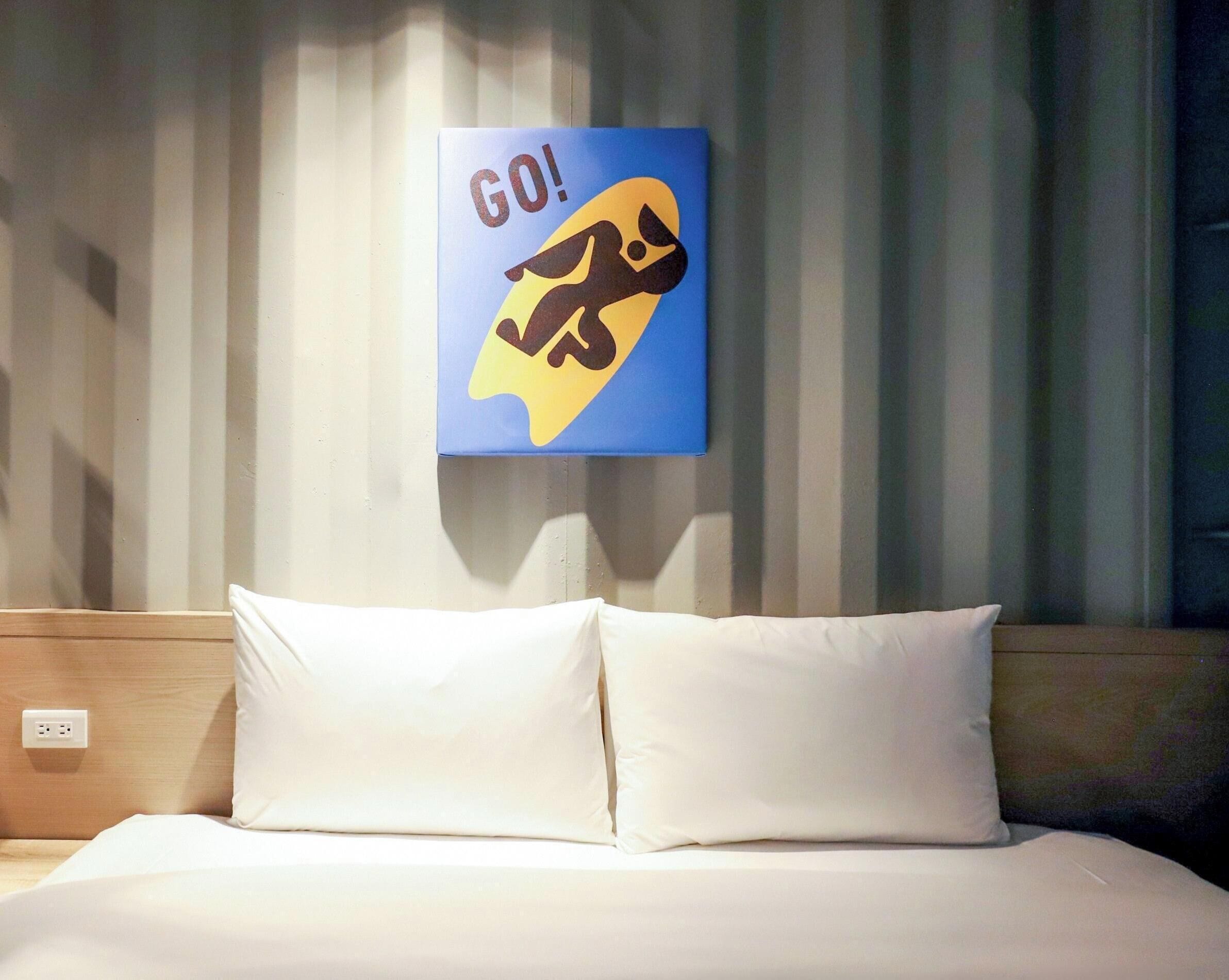 A hotel room with a bed, two pillows, and a wall-mounted sign with a graphic and the word "GO!"