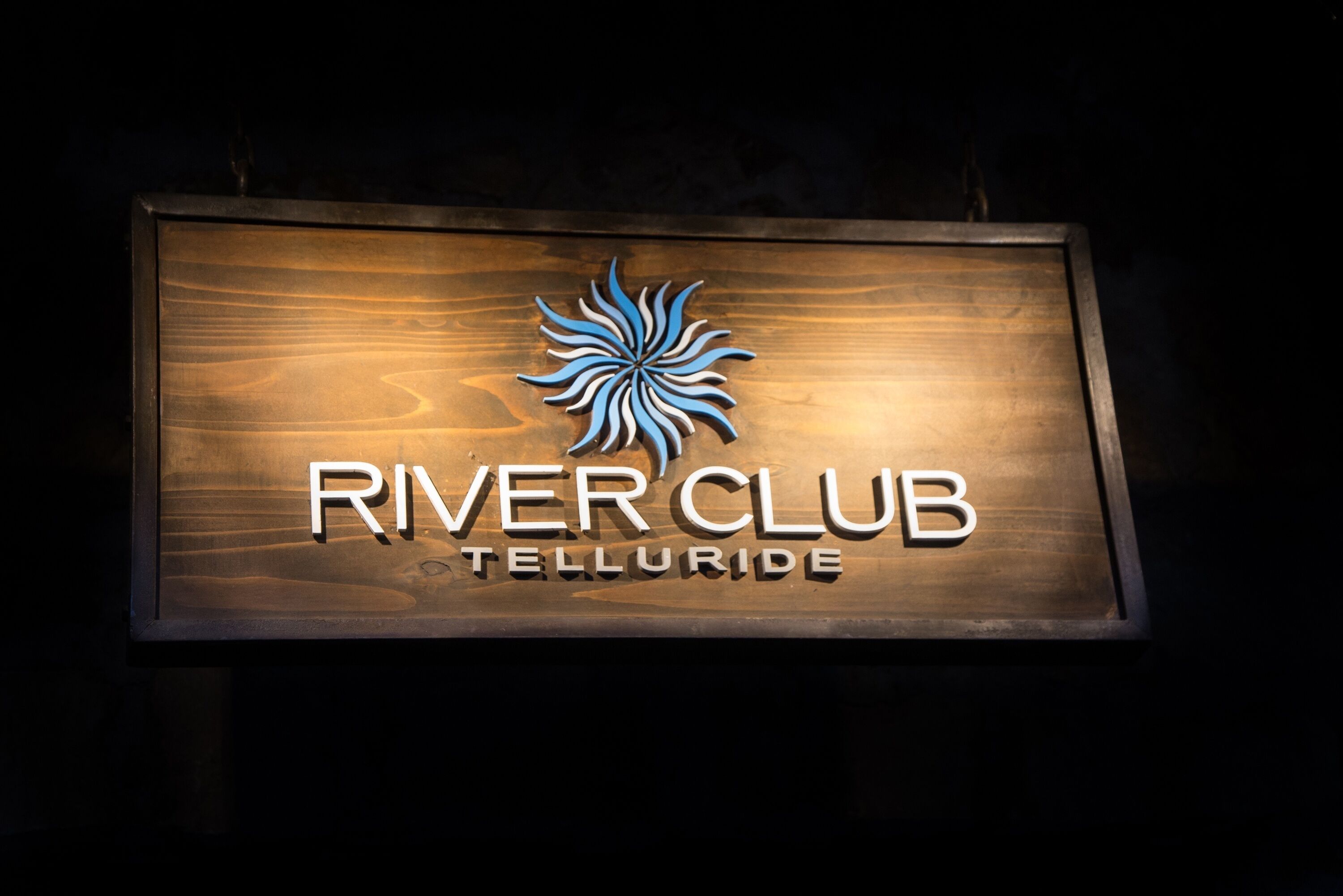 Ein hölzernes Schild mit dem Schriftzug 'River Club Telluride' und einem stilisierten Sonnenlogo.