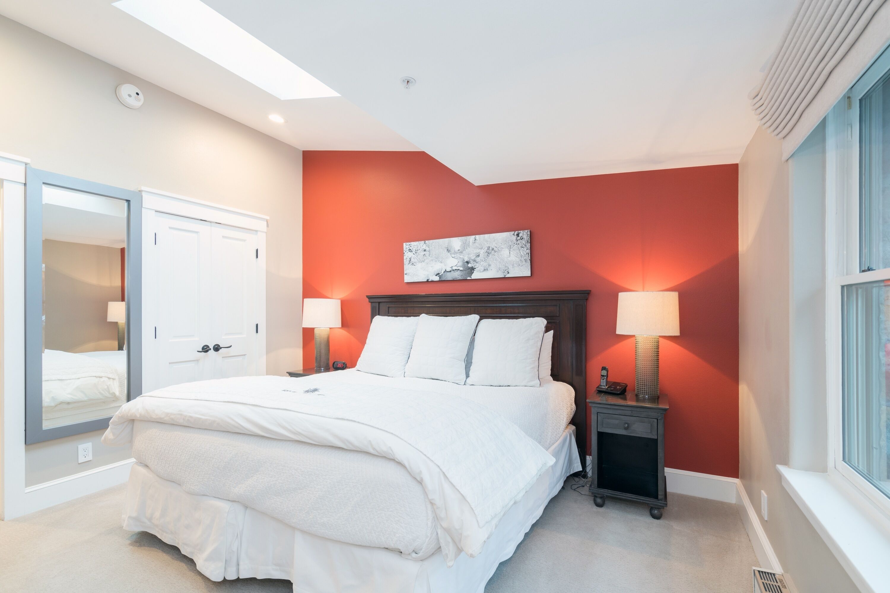 A bedroom with a large bed, a nightstand with a lamp, a mirror, and a window with blinds.