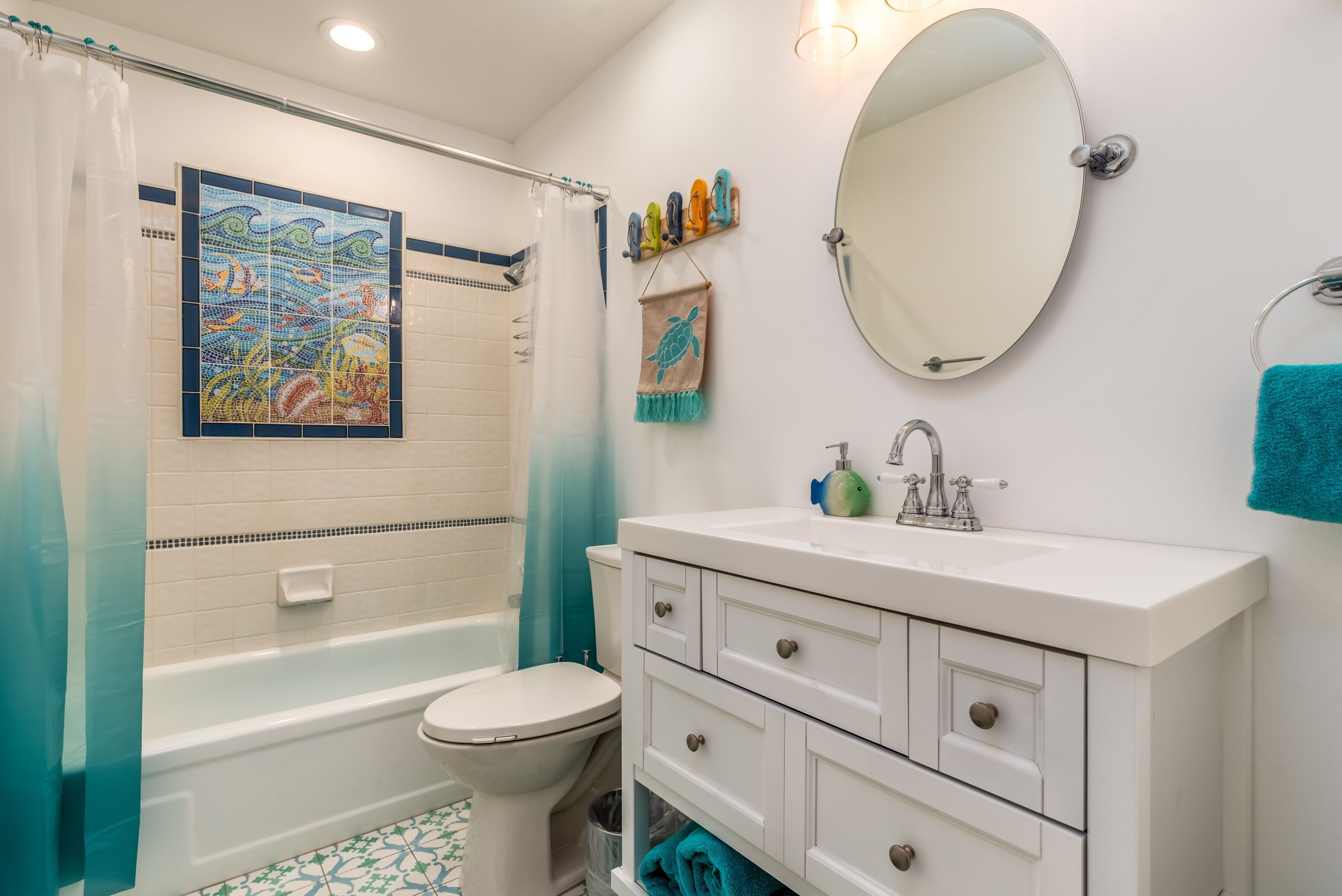 Un baño con bañera, inodoro, tocador blanco con cajones, un espejo y una obra de arte mural de mosaico.