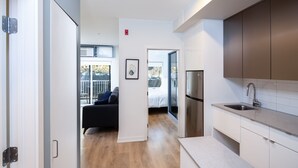 Een moderne keuken met een roestvrijstalen koelkast, een spoelbak en een aanrecht. Er ligt een houten vloer en in de aangrenzende woonkamer staat een zwarte bank.