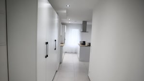Ein schmaler, gut beleuchteter Gang mit weißen Wänden und Fliesenboden, der zu einem Küchenbereich mit Fenster und Vorhängen führt.