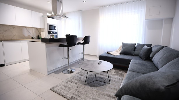 Ein modernes Wohnzimmer mit grauem Sofa, einem runden Couchtisch und einer Küchenzeile mit weißen Schränken und einer Marmor-Spritzwand.