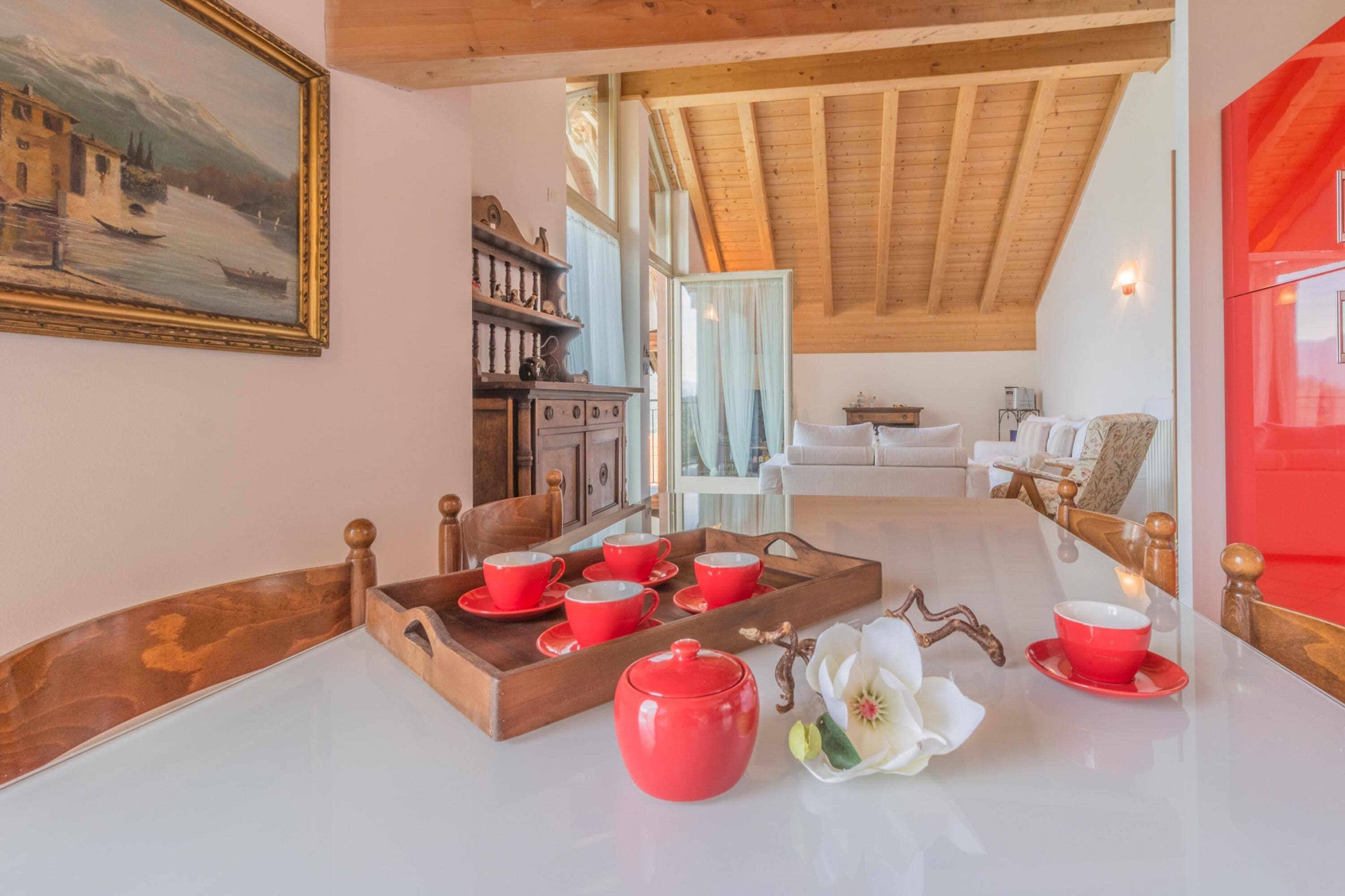 Una stanza accogliente con soffitto in legno, un quadro appeso al muro e un'area pranzo con tazze e piattini rossi.