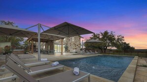 Una piscina con sillas de descanso y sombrillas, rodeada de muros de piedra y vistas al atardecer.