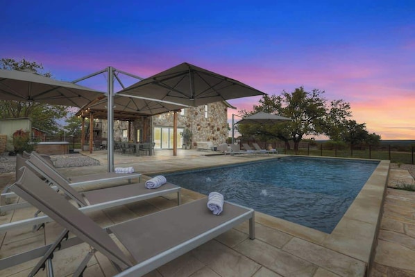 A swimming pool with lounge chairs and umbrellas, surrounded by stone walls and a sunset view.