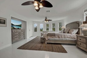 A bedroom with a large bed, a dresser, a nightstand, a ceiling fan, and a view of a lake through a window.