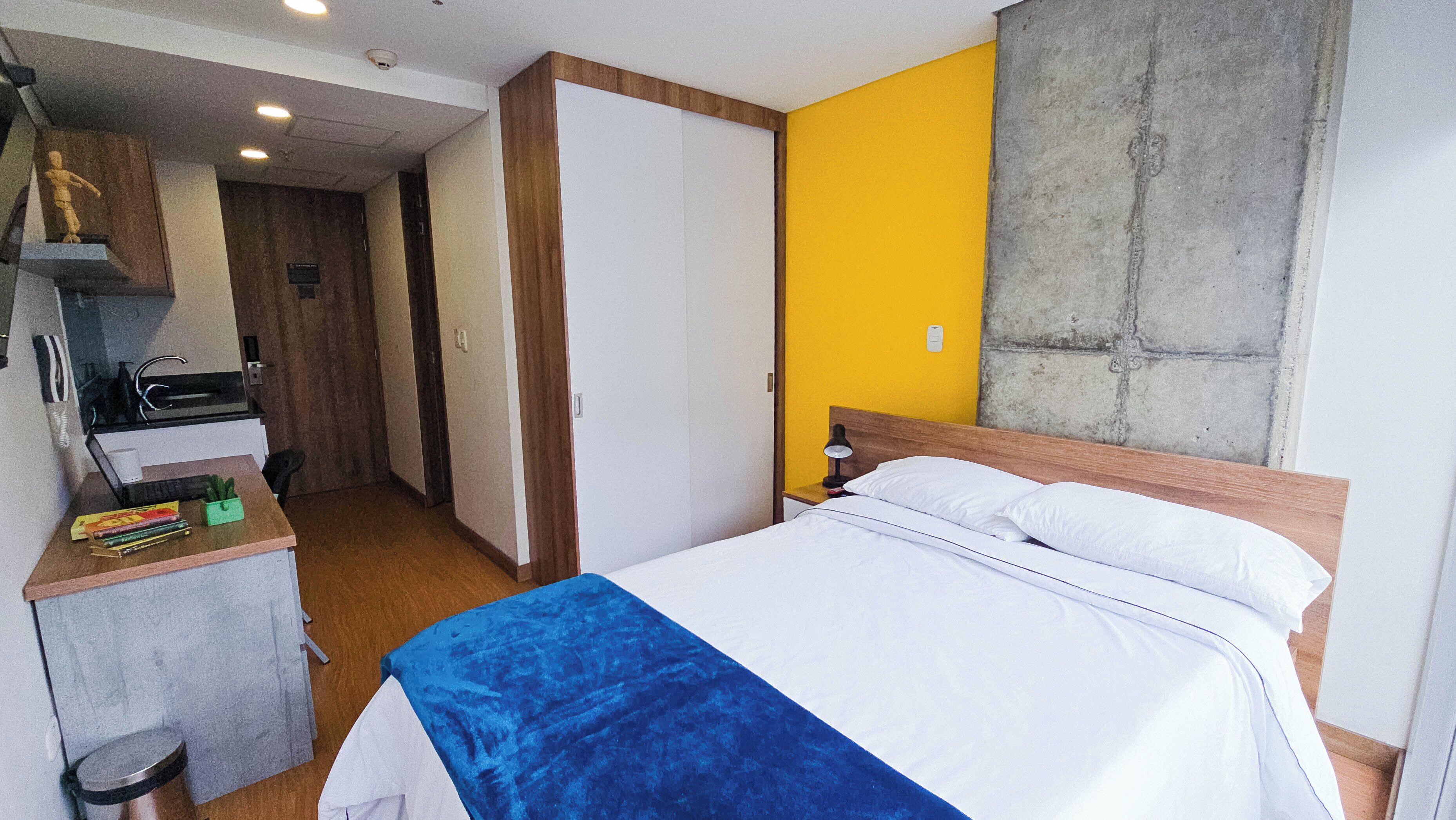 A hotel room with a bed, a desk with a chair, a small kitchenette, and a bathroom.