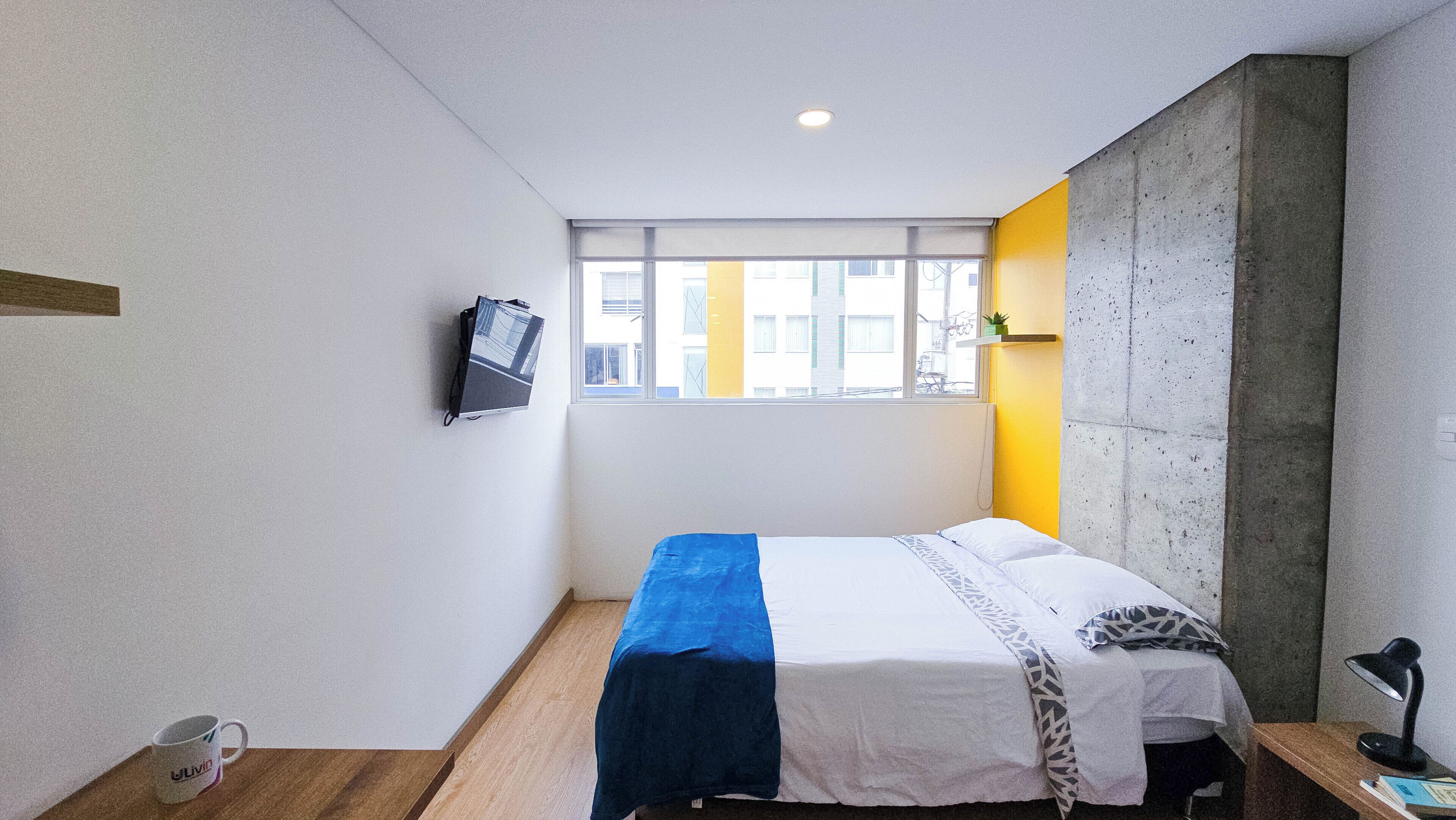 Habitación de hotel compacta con cama, televisor montado en la pared, ventana con vistas a edificios y una pared de concreto decorativa.