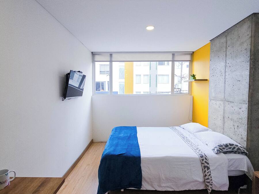 Habitación de hotel compacta con cama, televisor montado en la pared, ventana con vistas a edificios y una pared de concreto decorativa.