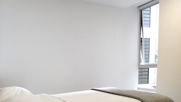 A single bed with a wooden headboard, a window with a view of buildings, and a ceiling light. - ULIVIN SAS (Manizales)