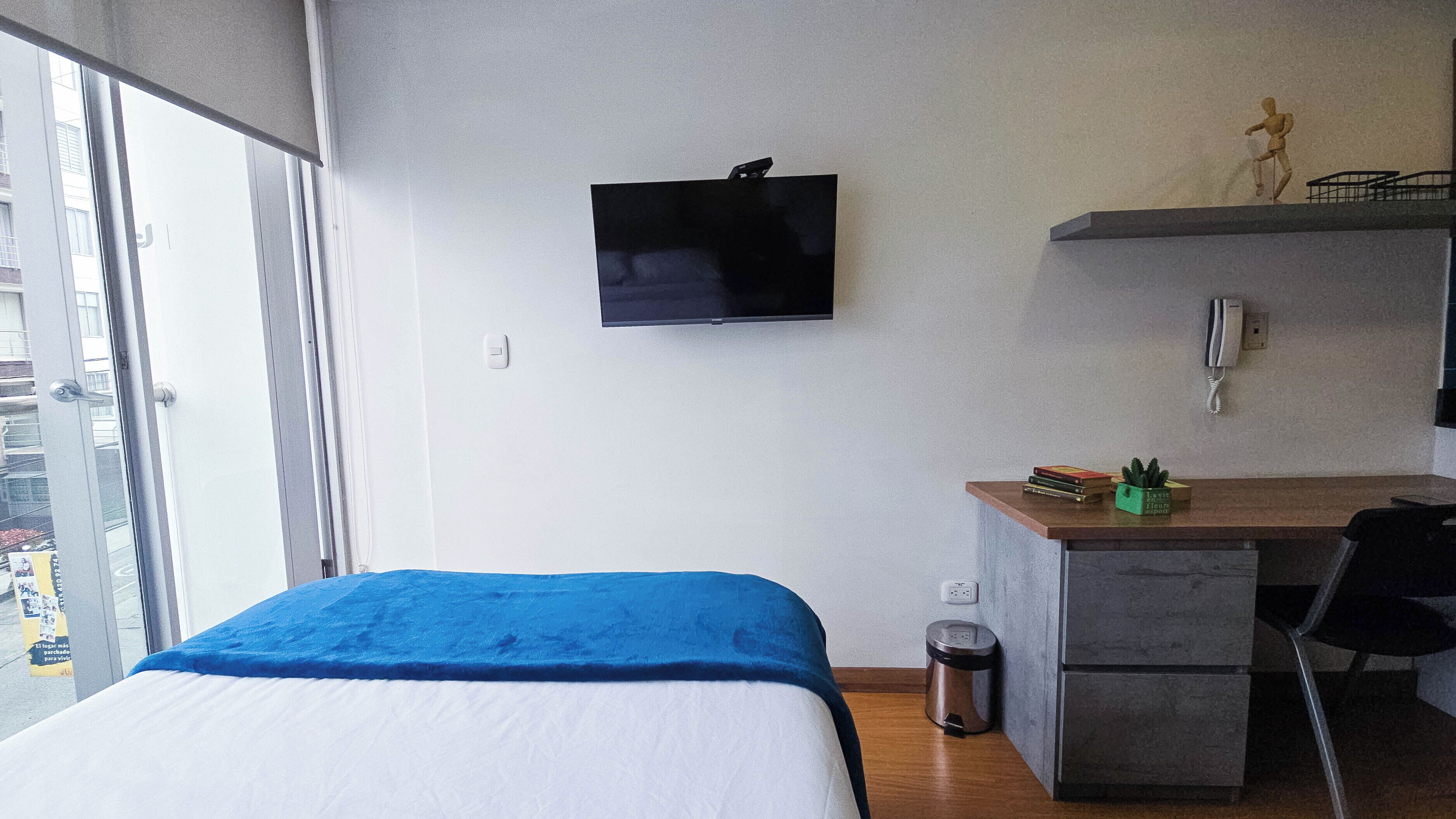 Habitación de hotel con cama, escritorio, cubo de basura, televisión y una pequeña estantería con una estatua.