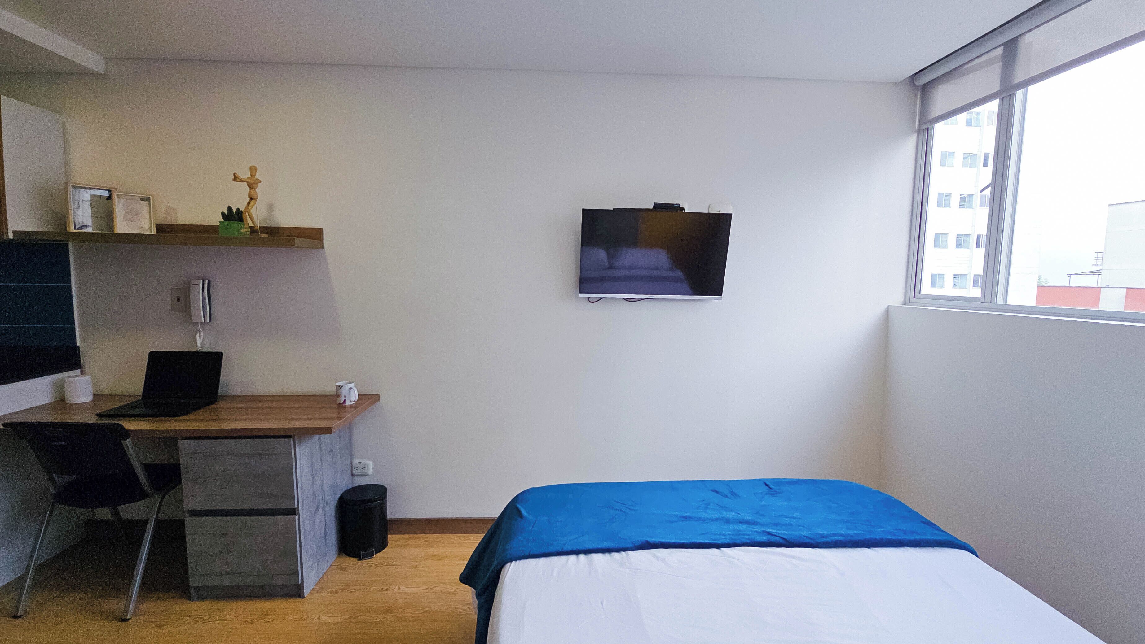 Une petite chambre d’hôtel moderne, équipée d’un lit, d’un bureau avec un ordinateur portable, d’une chaise, d’une petite poubelle et d’un téléviseur fixé au mur.