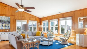 A spacious living room with wooden paneling, a ceiling fan, a large window, and a blue rug. - Adjust Your Focus, Almost Oceanfront Duplex in Emerald Isle, NC! (Emerald Isle)