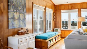 A cozy room with wooden walls, a large window with blinds, a bookshelf, and a blue cushioned bench. - Adjust Your Focus, Almost Oceanfront Duplex in Emerald Isle, NC! (Emerald Isle)