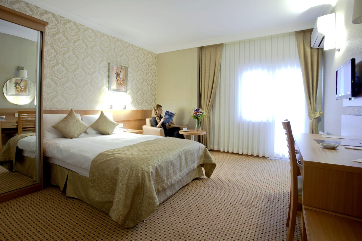 Habitación de hotel con cama, escritorio, silla, televisor, lámpara y ventana con cortinas.