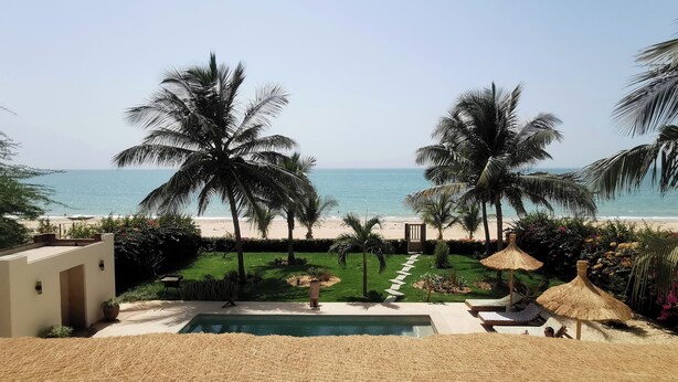 A pool area with a view of the beach, palm trees, and a thatched umbrella.