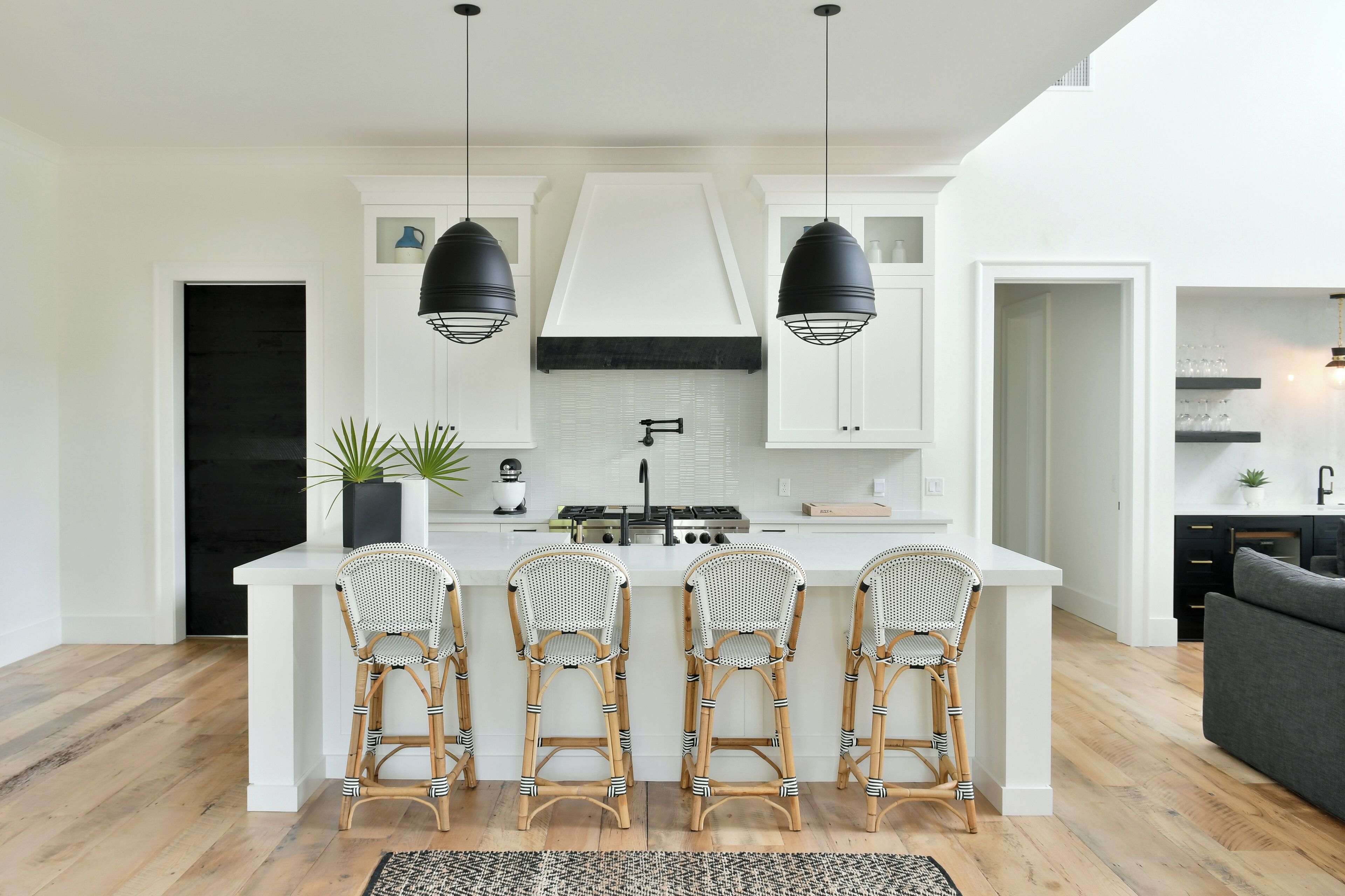 A modern kitchen with a central island, white cabinetry, black pendant lights, and three wicker bar stools.
