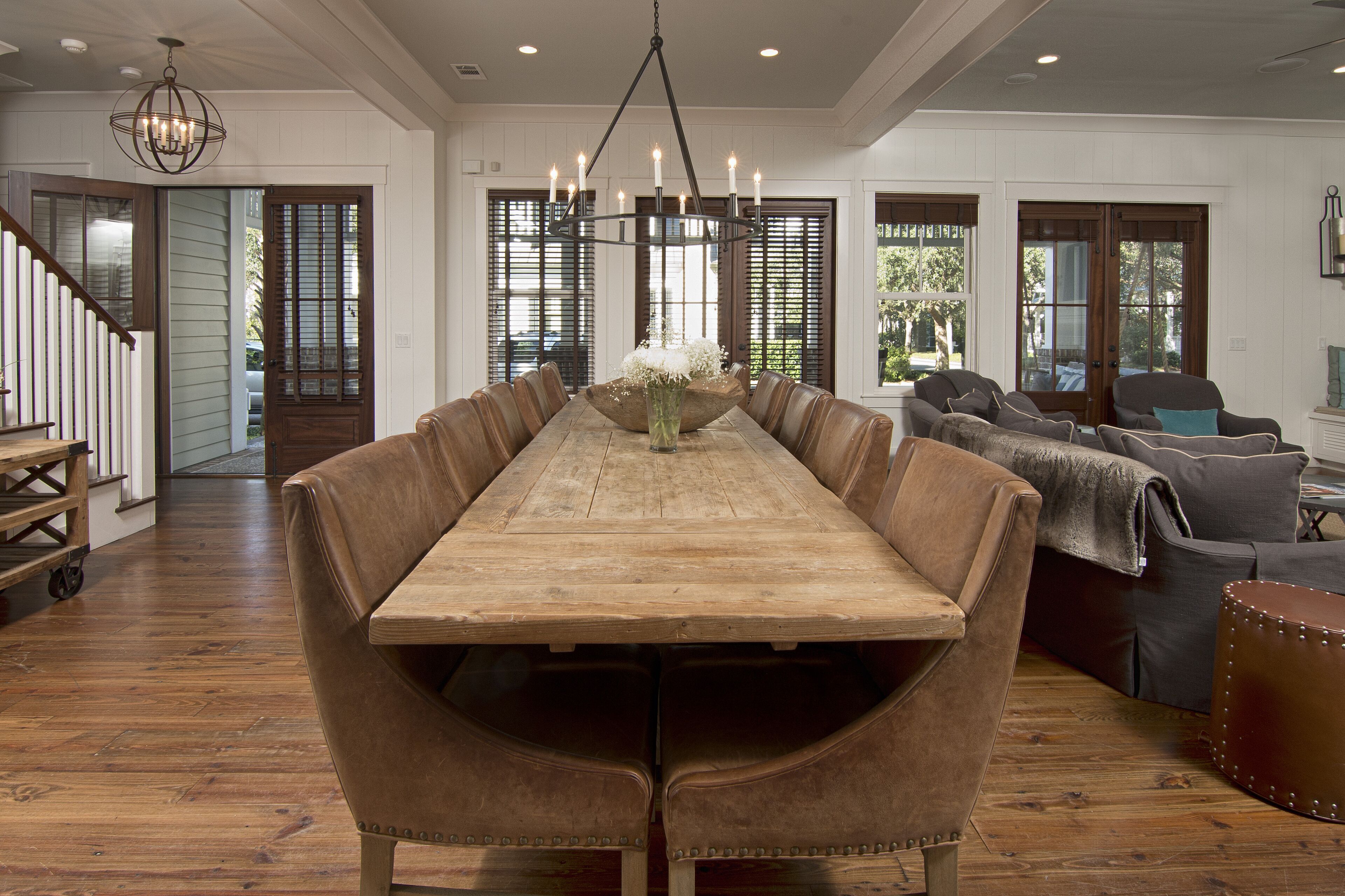 A dining area with a long wooden table and leather chairs, a chandelier, and a view of the outdoors through large windows.