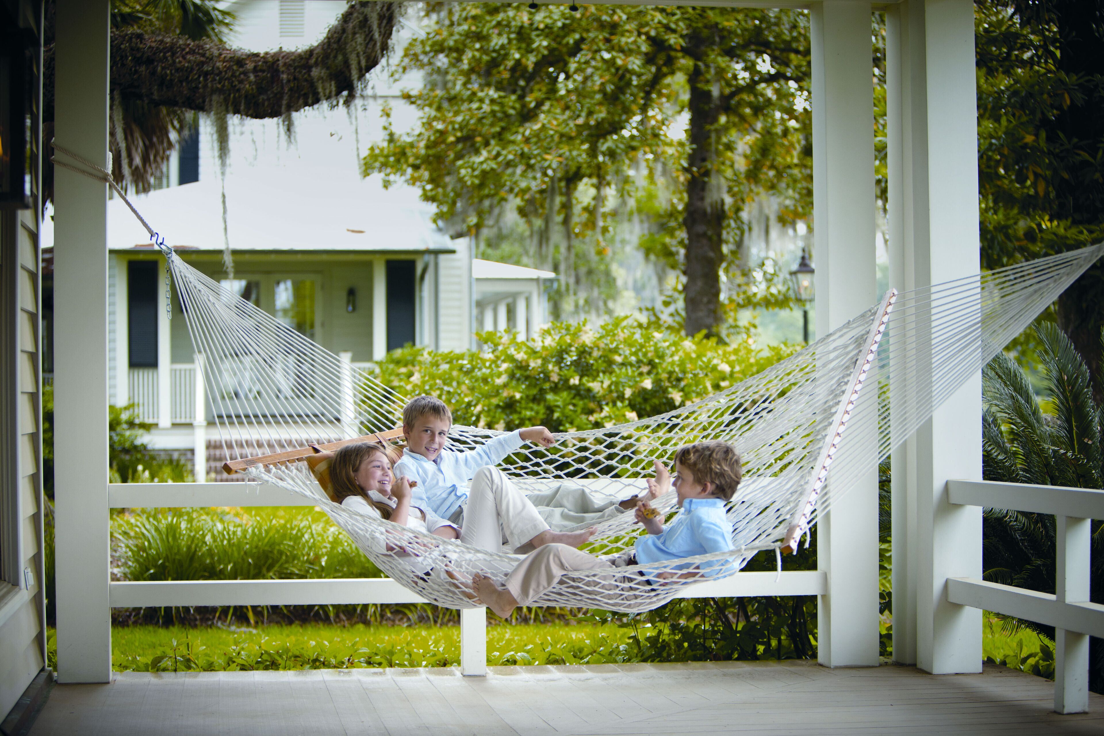 A hammock on a porch with a view of a house and greenery.