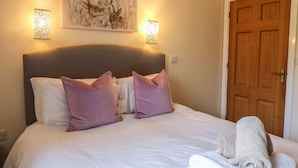 A neatly made bed with white and purple pillows, a headboard, and a painting above it. There are two wall-mounted lamps on either side of the bed. - Caban Dulas (Rhayader)