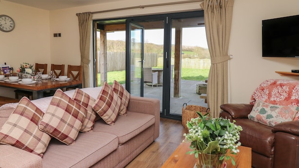 A living room with a sofa, a coffee table with a floral arrangement, a television, and a dining area with a table set for four. - Caban Dulas (Rhayader)