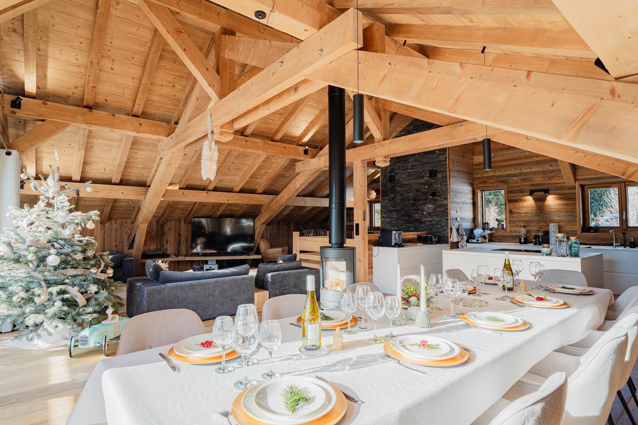 A dining area with a long table set for a meal, a Christmas tree, and a kitchen in the background.