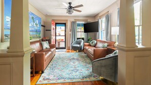 A spacious living room with a large window, a ceiling fan, and a colorful rug.