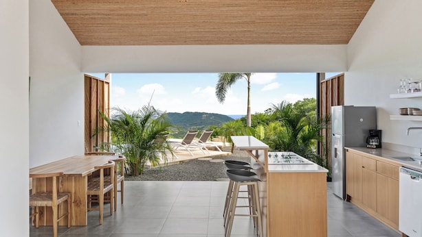 A modern kitchen with a bar area, a dining table, and chairs. There is a view of a patio with chairs and plants.